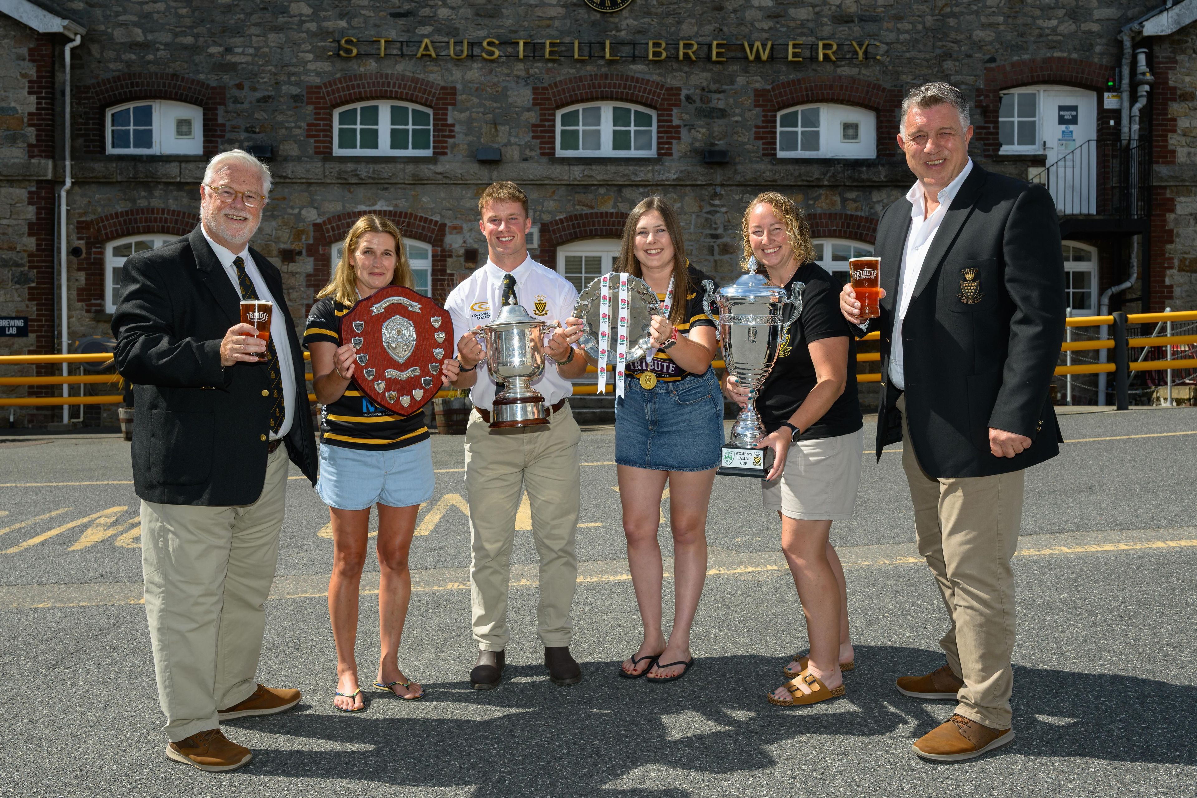 St Austell Brewery team members pose with players and representatives from Cornwall's Mens and Womens rugby teams, holding trophies with the St Austell Brewery sign in the background