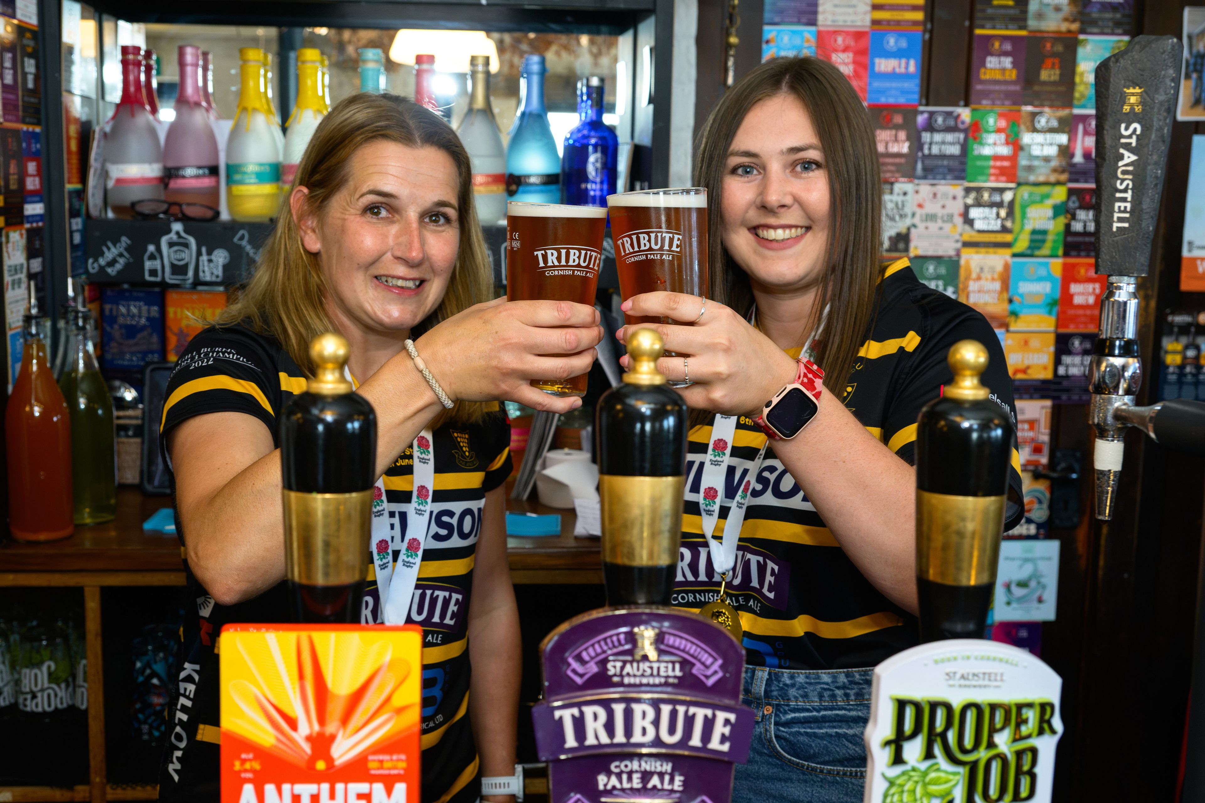 Two members of Cornwall Women's Rugby team cheers two pints of Tribute whilst stood behind a bar
